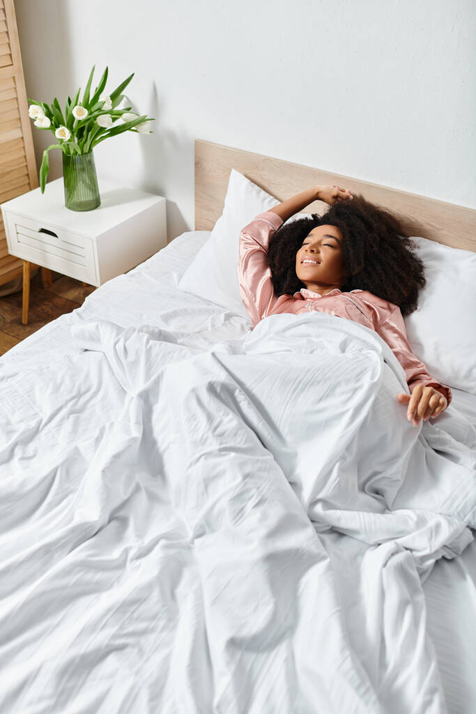 Femme afro-américaine bouclée en pyjama couchée sur un lit avec des draps blancs, profitant d'une matinée paisible. - Photo, image