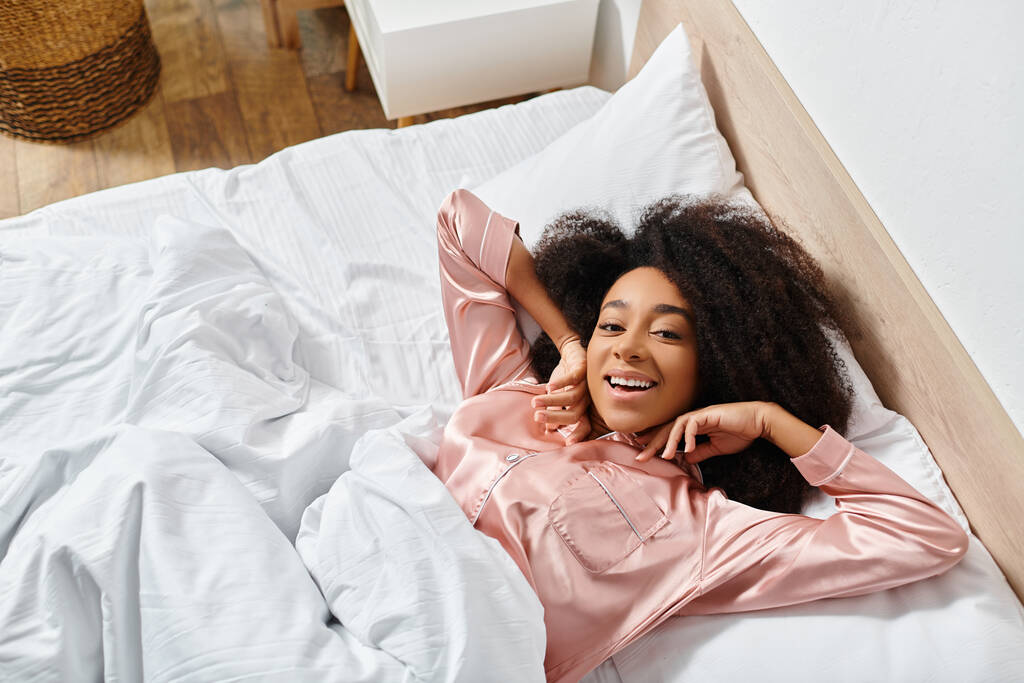 Femme afro-américaine frisée en pyjama reposant paisiblement sur un lit blanc dans une chambre sereine le matin. - Photo, image