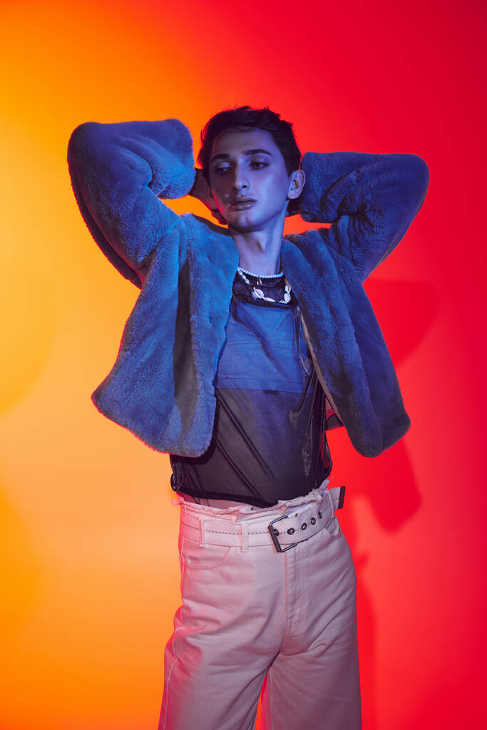 attraktiver modisch androgyner Mann in lila Kunstpelz posiert vor lebendiger Kulisse und schaut weg - Foto, Bild