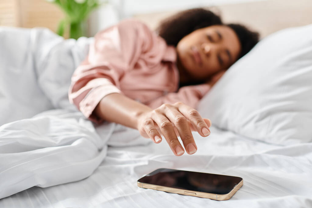 Mujer afroamericana rizada en pijama relajándose en la cama, absorta en su teléfono celular por la mañana. - Foto, imagen