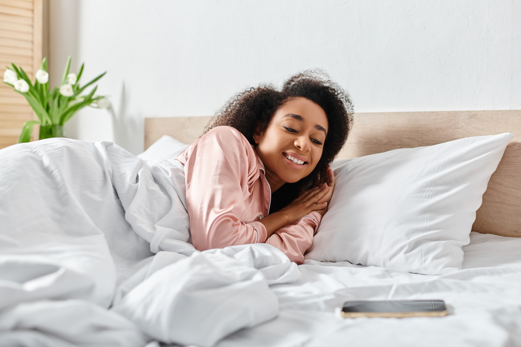 Pijamalı kıvırcık Afrikalı Amerikalı kadın rahat bir yatak odasında beyaz çarşaflı bir yatakta uzanıyor.. - Fotoğraf, Görsel