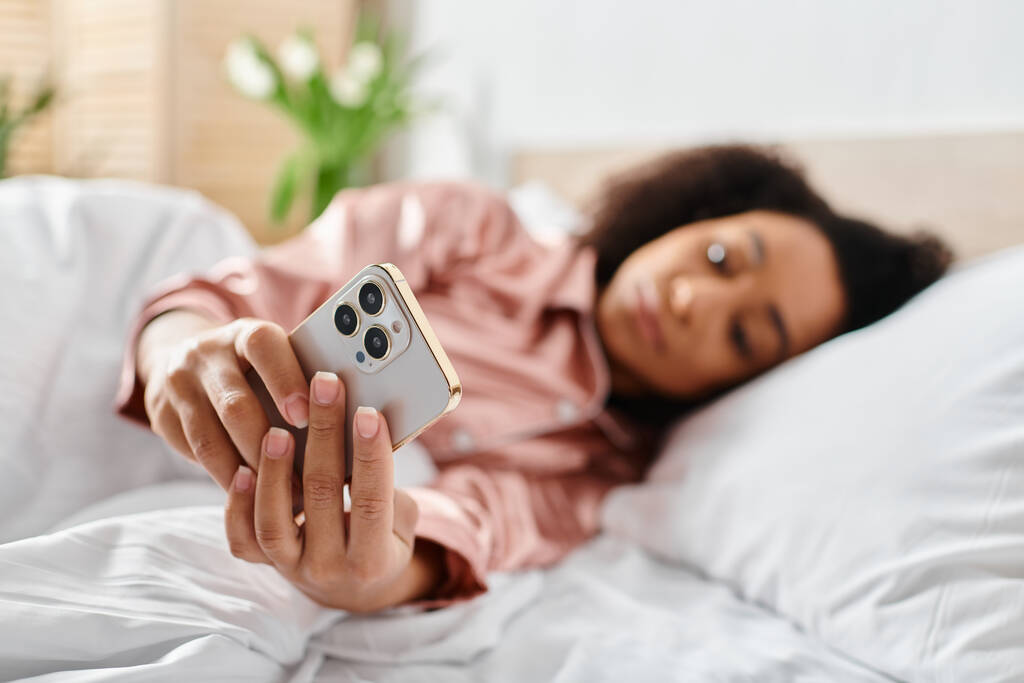 Σγουρή Αφρο-Αμερικανίδα με πιτζάμες ξαπλωμένη στο κρεβάτι, κρατώντας ένα τηλεχειριστήριο στο χέρι της, χαλαρώνοντας το πρωί. - Φωτογραφία, εικόνα