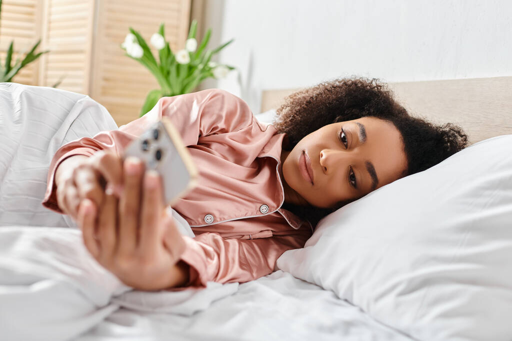 Kudrnatá Afroameričanka v pyžamu ležící v posteli, ráno pokojně drží dálkové ovládání. - Fotografie, Obrázek