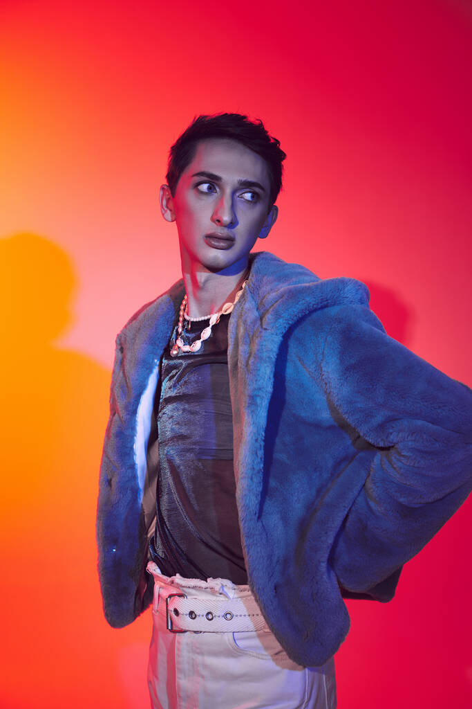attractive modish androgynous man in purple faux fur posing on vibrant backdrop and looking away - Photo, Image