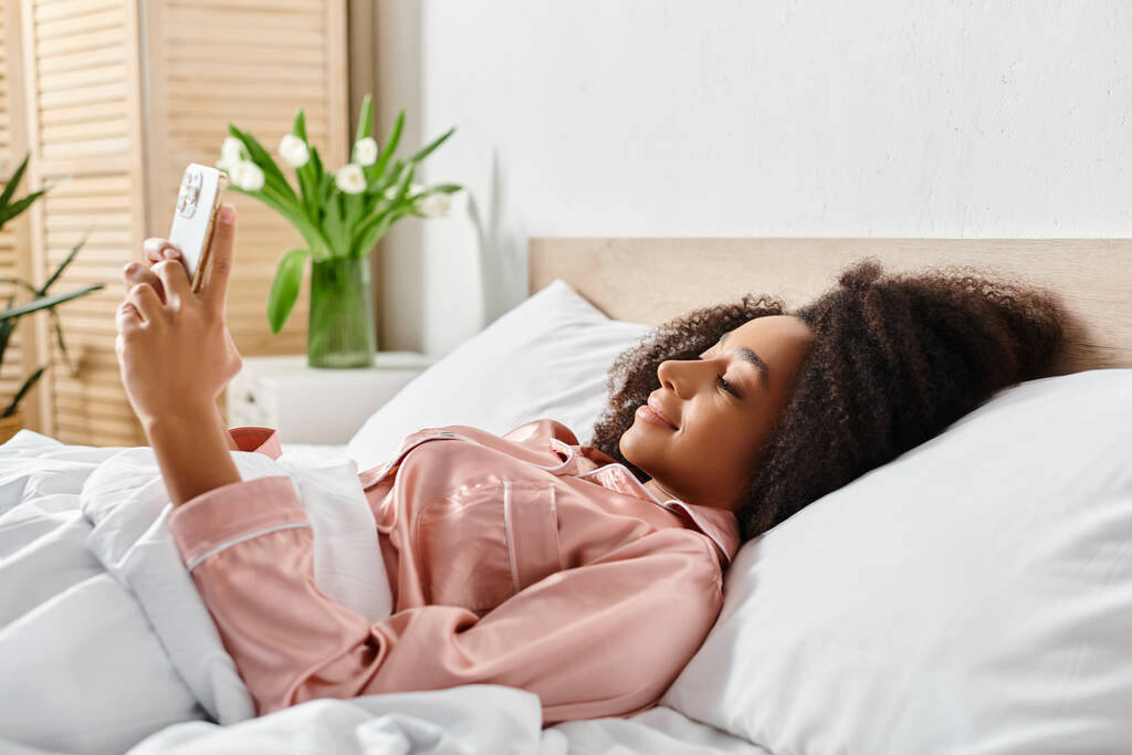 パジャマのカーリー・アフリカ系アメリカ人女性がベッドで寝て携帯電話を持っている - 写真・画像