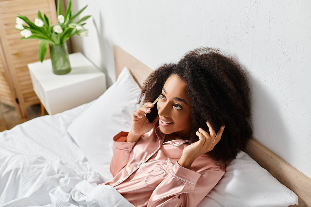 Kręcone Afroamerykanka kobieta w piżamie rozmawia przez telefon komórkowy podczas leżąc na łóżku w sypialni rano. - Zdjęcie, obraz