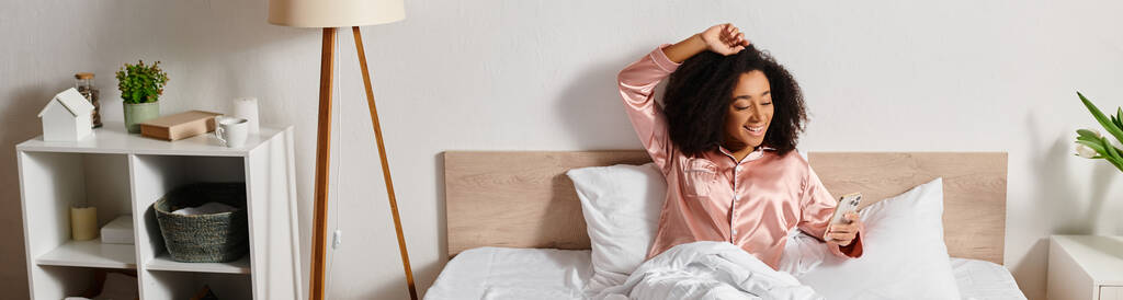 Kıvırcık Afrikalı Amerikalı bir kadın yatak odasındaki bir yatağın üzerinde dinleniyor, sakin bir sabah boyunca pembe bir gömlek giyiyor.. - Fotoğraf, Görsel