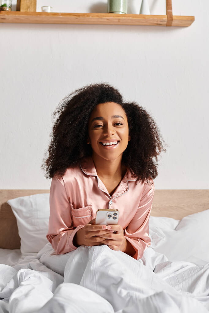 Σγουρή Αφρο-Αμερικανίδα γυναίκα με πιτζάμες κάθεται στο κρεβάτι, απορροφημένη στην οθόνη του κινητού της το πρωί. - Φωτογραφία, εικόνα