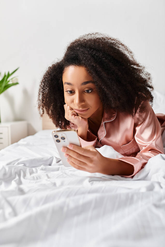 Curly African American woman in pajamas lying on bed, engrossed in tablet at morning time. - Photo, Image