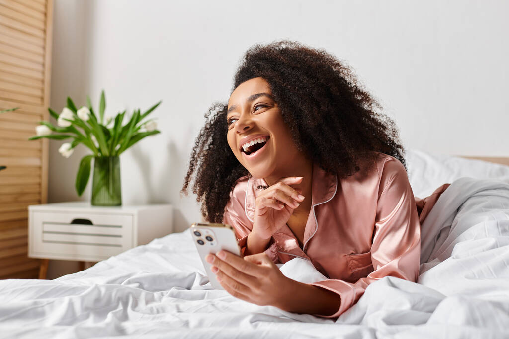 Una mujer afroamericana rizada, vestida en pijama, descansando pacíficamente en una cama mientras sostiene un teléfono celular. - Foto, Imagen