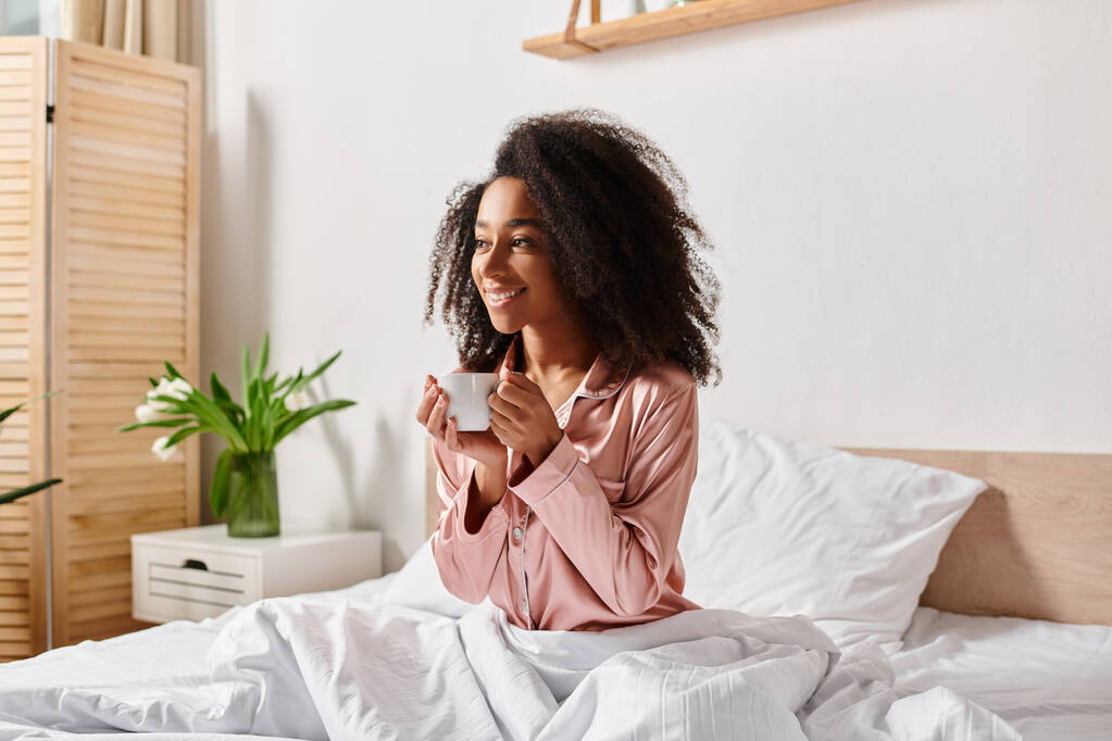 Una mujer afroamericana rizada en pijama sentada en una cama, disfrutando de una taza de café a la luz del sol de la mañana. - Foto, Imagen