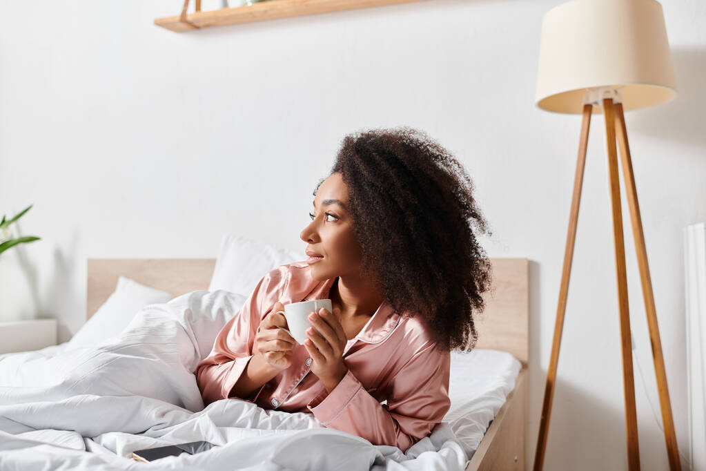 Kręcone Afroamerykanka w piżamie relaksuje się w łóżku z filiżanką kawy w godzinach porannych. - Zdjęcie, obraz