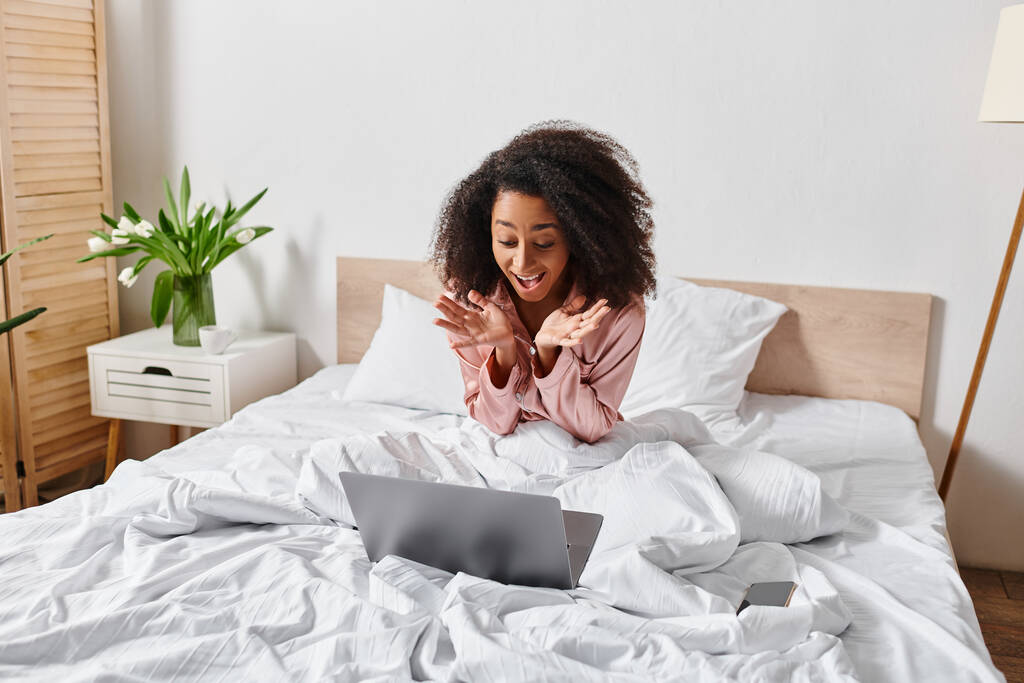 Curly African American woman in pajamas, sitting on bed in bedroom, absorbed in using laptop in morning time. - Photo, Image