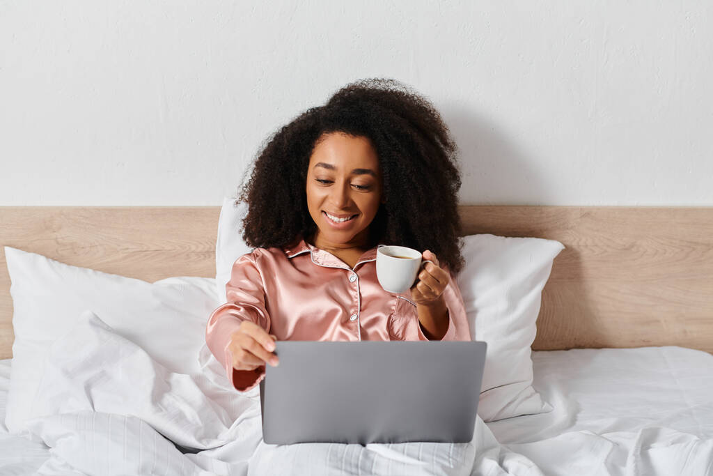 Curly mulher afro-americana de pijama sentado na cama, segurando uma xícara de café, olhando para a tela do laptop no quarto. - Foto, Imagem