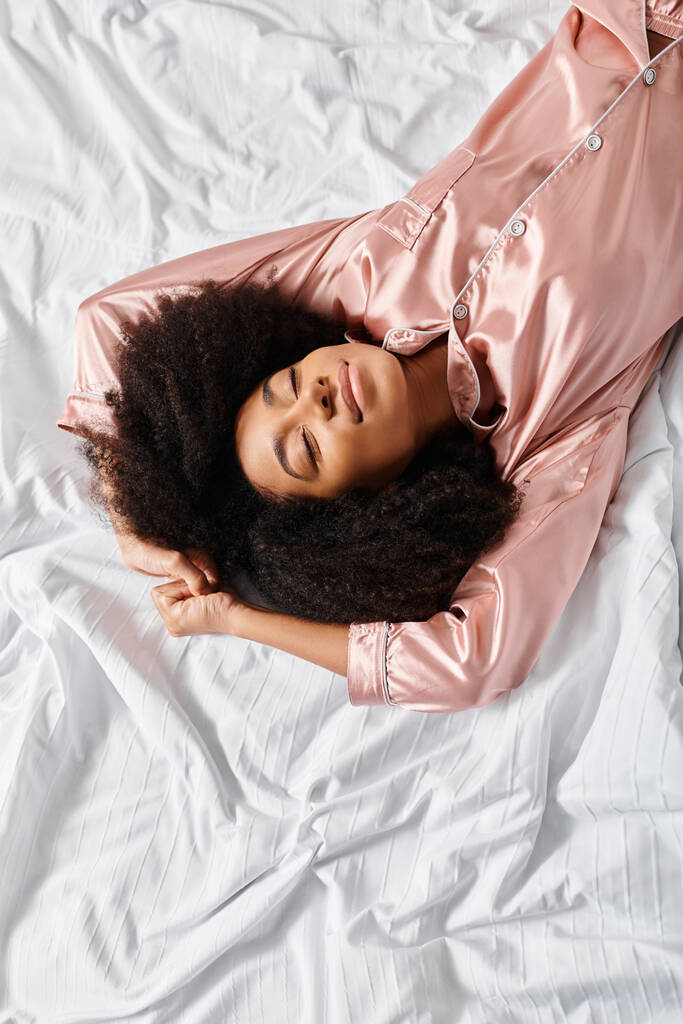 パジャマに住むカーリーなアフリカ系アメリカ人女性が,寝室の白いベッドの上に平和的に横たわっている. 朝の時間です. - 写真・画像