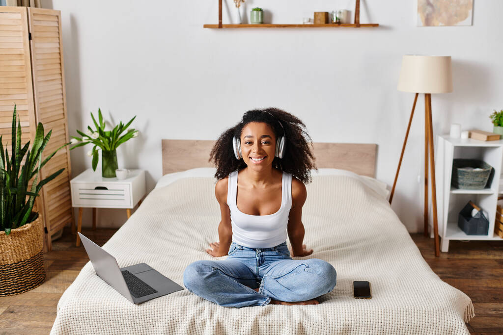 現代の寝室のラップトップスクリーンに焦点を当てたベッドの上に座っているタンクのカーリーアフリカ系アメリカ人女性. - 写真・画像