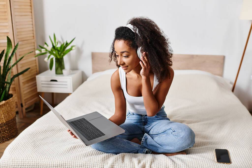 タンクの上のカーリーアフリカ系アメリカ人女性はスタイリッシュなモダンなベッドルームでラップトップコンピュータを使用してベッドに座ります. - 写真・画像