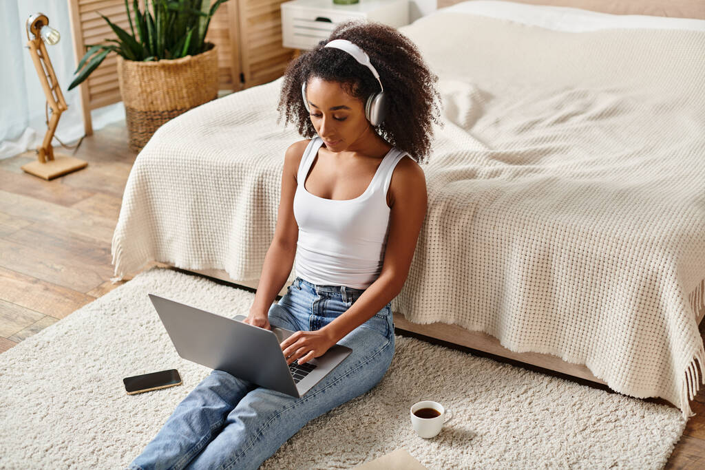 A curly African American woman in a tank top sits on the floor using a laptop in a modern bedroom. - Photo, Image