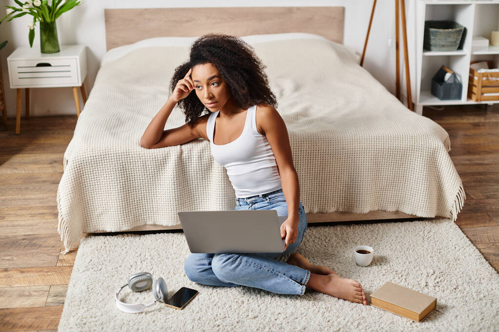 Una mujer afroamericana rizada en una camiseta sin mangas se sienta en el suelo, absorto en el uso de un ordenador portátil en un dormitorio moderno. - Foto, Imagen