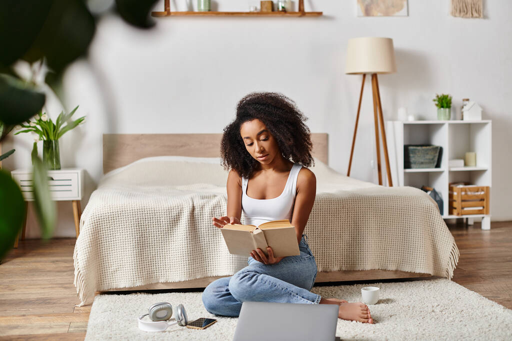 Askılı tişörtlü kıvırcık Afrikalı Amerikalı bir kadın, modern bir yatak odasında kitap okumaya dalmış.. - Fotoğraf, Görsel