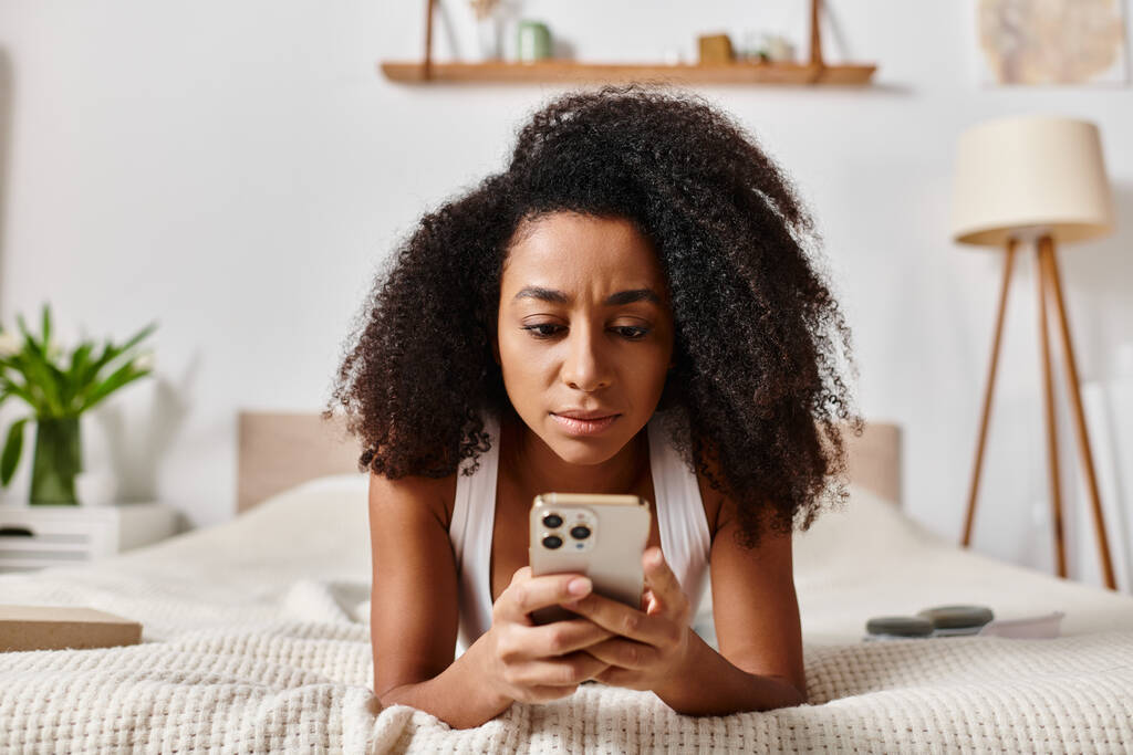 Kolsuz tişörtlü kıvırcık Afrikalı Amerikalı kadın, bir yatağa uzanmış, cep telefonuyla modern bir yatak odasına dalmış.. - Fotoğraf, Görsel