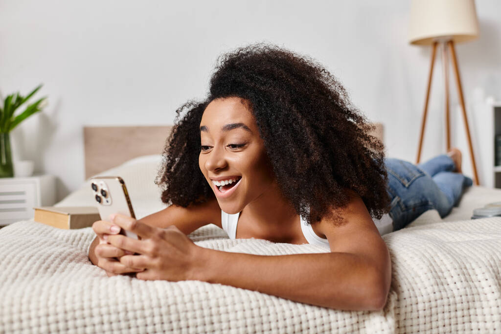 Una mujer afroamericana rizada en una camiseta sin mangas relajándose en una cama con teléfono - Foto, imagen
