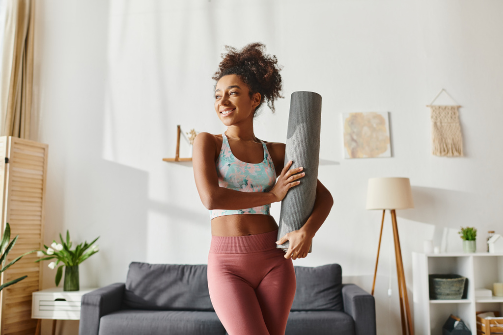 Una donna afroamericana riccia in abbigliamento attivo tiene un tappetino da yoga in un salotto elegante, pronto per la sua routine di allenamento. - Foto, immagini