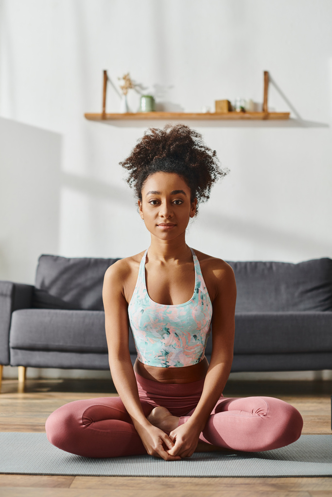 Femme afro-américaine bouclée en vêtements de sport faisant du yoga sur un tapis dans un cadre confortable salon. - Photo, image