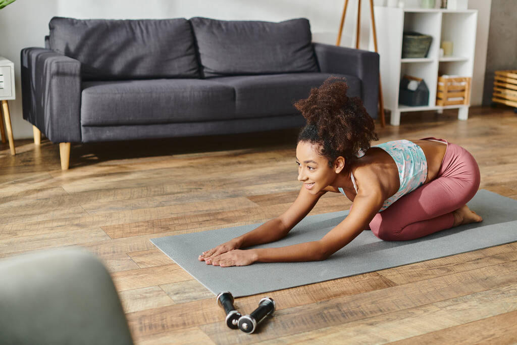 Mujer afroamericana rizada en desgaste activo con gracia sosteniendo una pose de yoga en una esterilla de yoga en casa. - Foto, Imagen