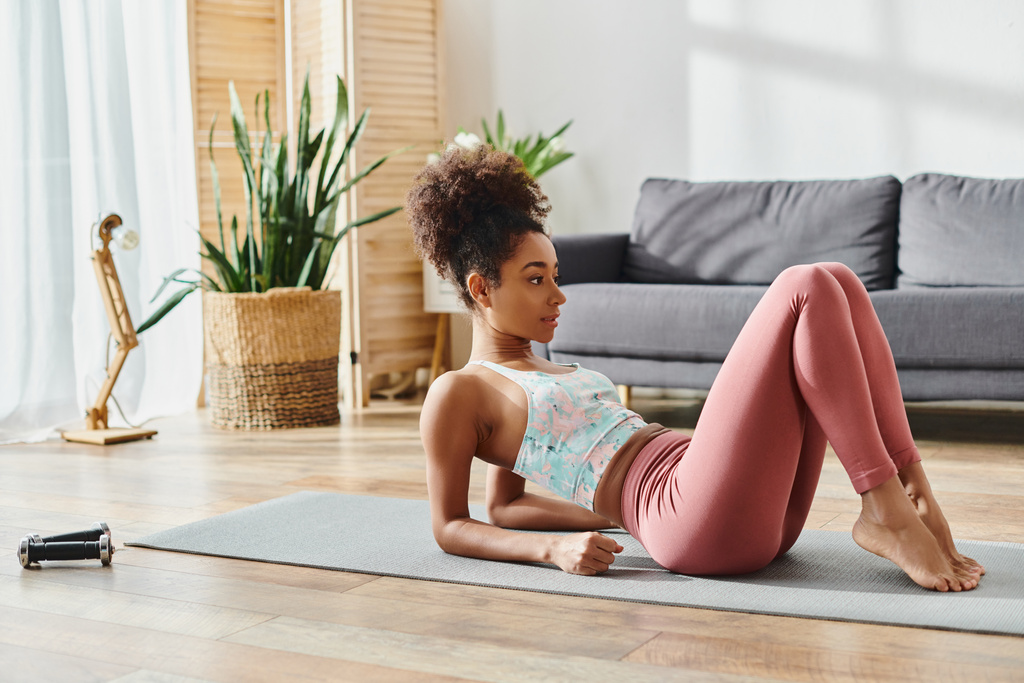Lockige Afroamerikanerin in aktiver Kleidung führt zu Hause anmutig eine Yoga-Pose auf einer bunten Yogamatte aus. - Foto, Bild