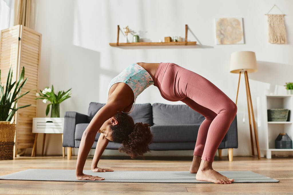 krullend Afro-Amerikaanse vrouw in actieve dragen sierlijk voert een handstand op een yoga mat in een serene home setting. - Foto, afbeelding