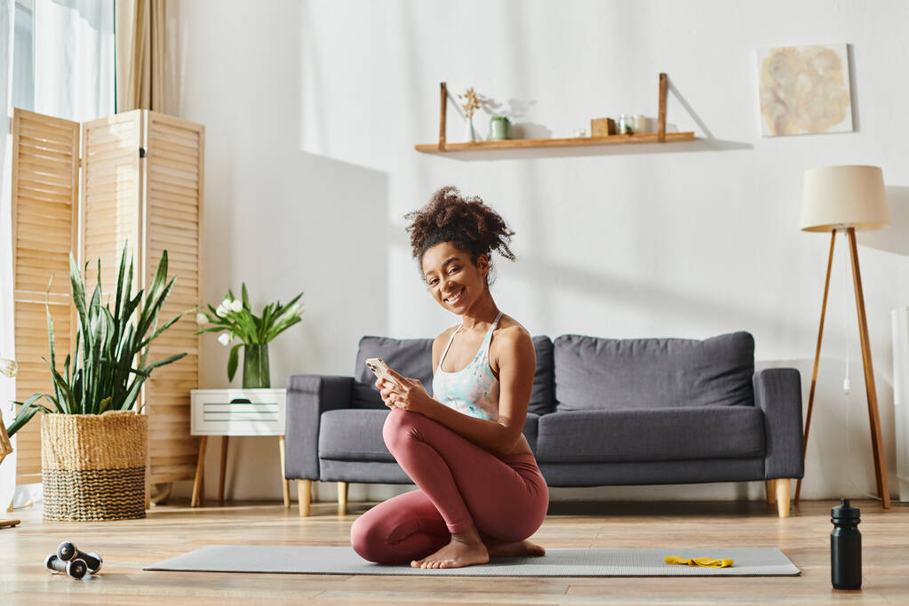 Una mujer afroamericana rizada en ropa deportiva sentada en una esterilla de yoga, absorta en la pantalla de su teléfono mientras hace ejercicio en casa. - Foto, imagen