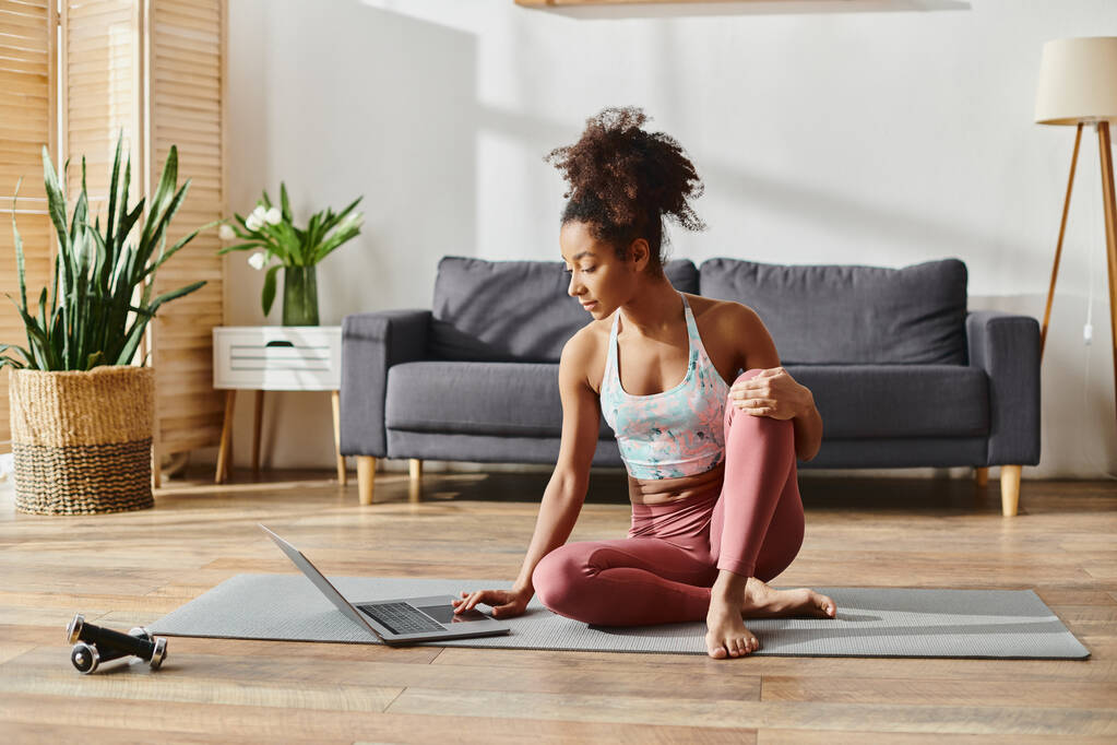 Eine lockige Afroamerikanerin in aktiver Kleidung sitzt friedlich auf einer Yogamatte in einem gemütlichen Wohnzimmer. - Foto, Bild