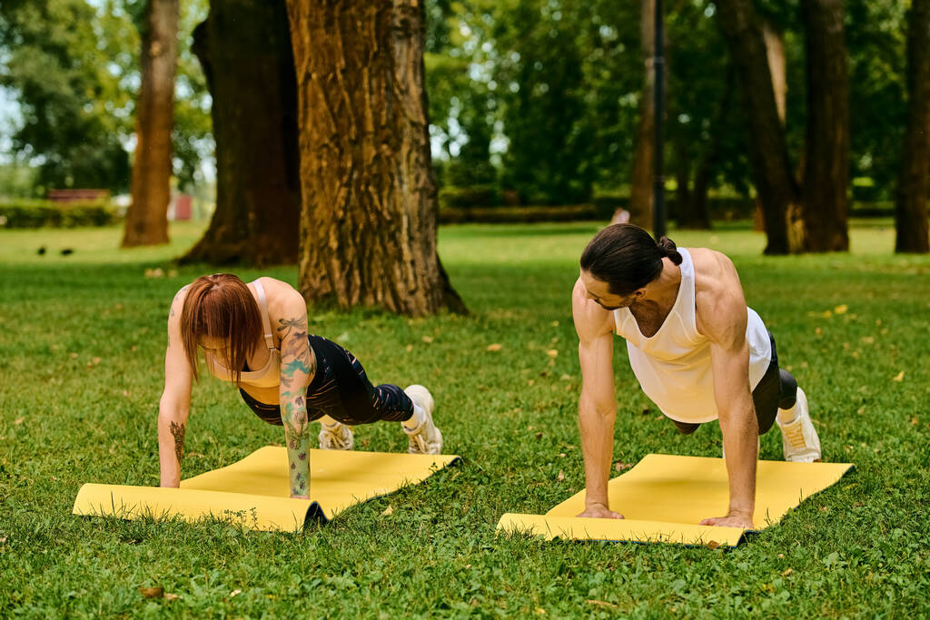 Un hombre y una mujer en ropa deportiva demostrando fuerza y determinación mientras realizan flexiones en un parque - Foto, Imagen