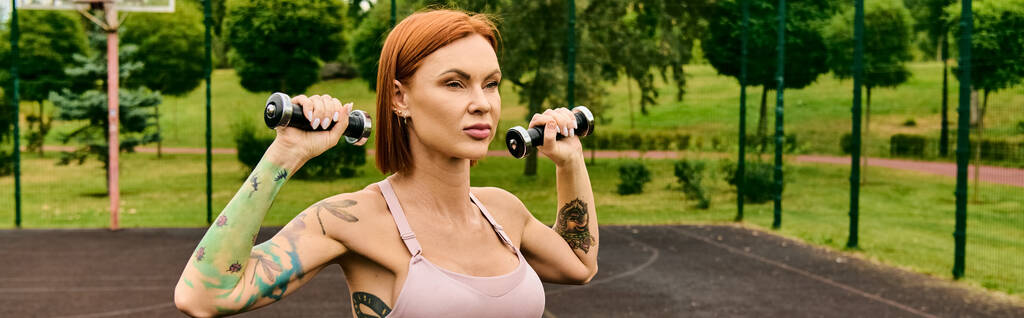 Egy határozott nő a parkban megragad két súlyzót, fókuszálva és eltökélten gyakorol. - Fotó, kép