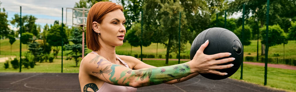 A woman in sportswear, holding a medicine ball, trains outdoors with determination - Photo, Image