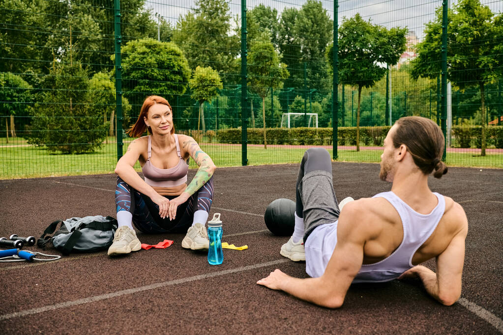 Ένας άνδρας και μια γυναίκα με αθλητικά κάθονται σε ένα γήπεδο μπάσκετ, μοιράζοντας αποφασιστικότητα και κίνητρα καθώς γυμνάζονται μαζί.. - Φωτογραφία, εικόνα
