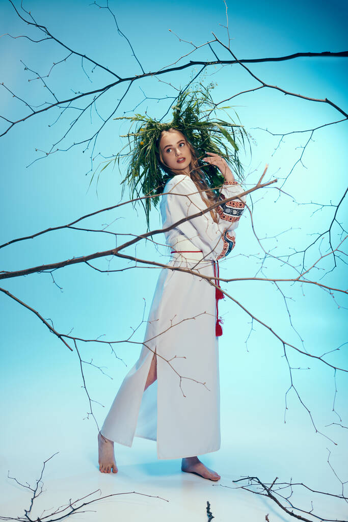 Un joven mavka en un vestido blanco con gracia sostiene una delicada rama en un entorno de estudio caprichoso. - Foto, imagen