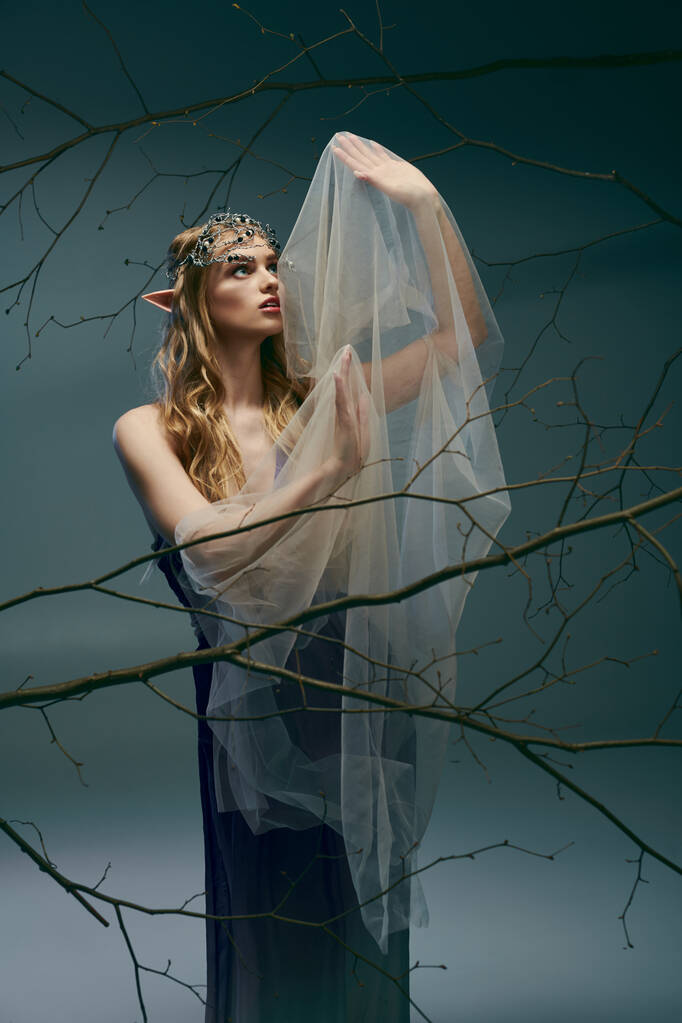 Μια νεαρή γυναίκα ντυμένη σαν πριγκίπισσα ξωτικό στέκεται με χάρη μπροστά σε ένα μεγαλοπρεπές δέντρο φορώντας πέπλο. - Φωτογραφία, εικόνα