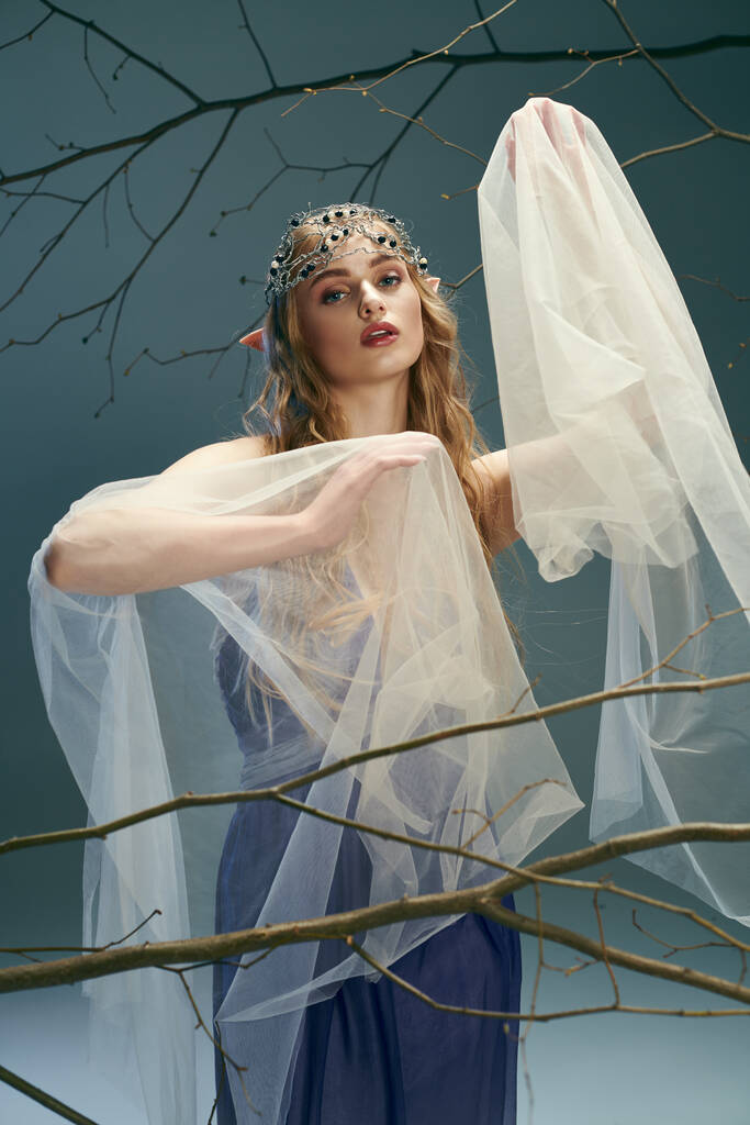 Młoda kobieta w pięknej niebieskiej sukience i płynącej białej welonie, ucieleśniająca istotę elfowej księżniczki z bajki lub fantazji w studio. - Zdjęcie, obraz