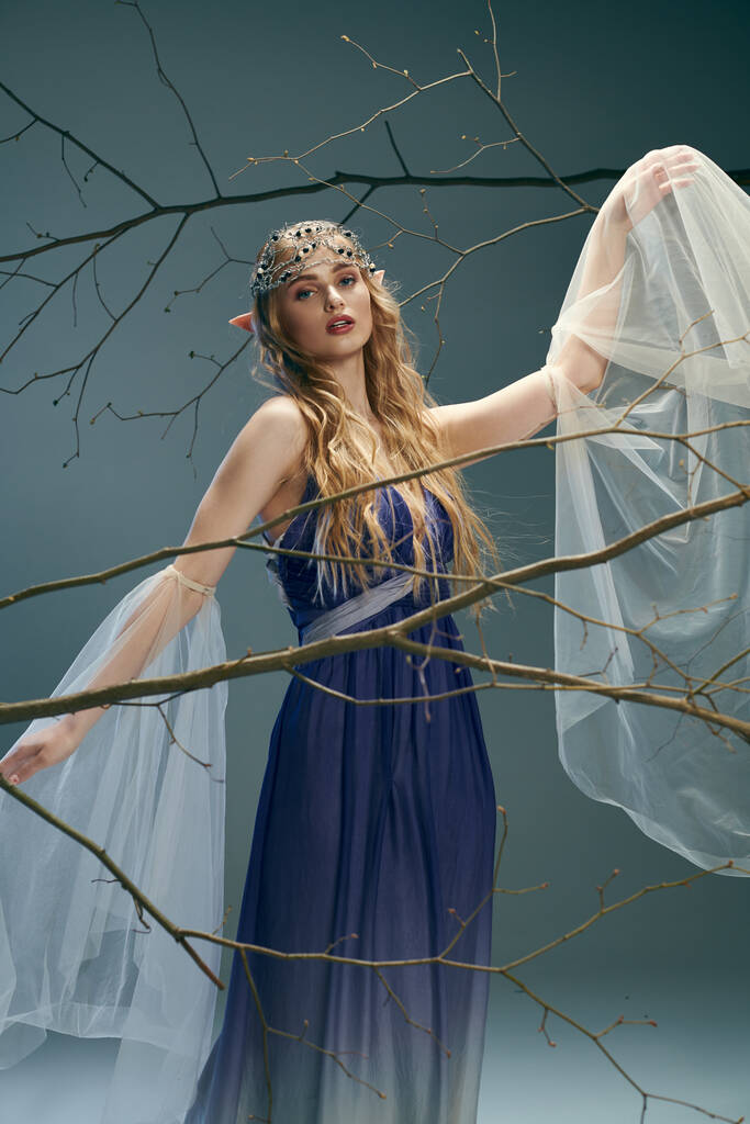 Eine junge Frau in einem blauen Kleid steht anmutig vor einem hoch aufragenden Baum und verkörpert eine ätherische Märchenprinzessin in einem Atelierambiente. - Foto, Bild