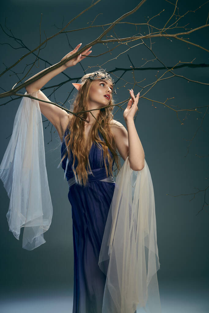 Młoda kobieta w niebieskiej sukience przypominającej elfową księżniczkę, delikatnie trzyma gałąź w studio. - Zdjęcie, obraz