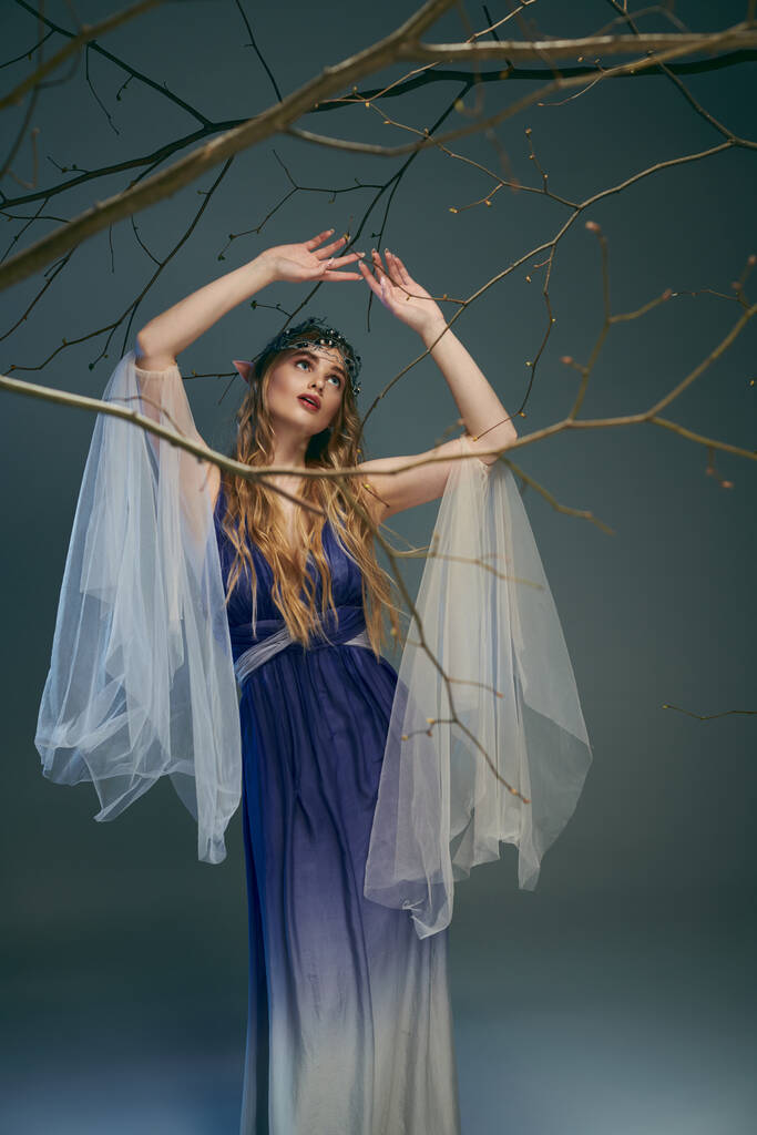 Una joven con un vestido azul y blanco se levanta con gracia junto a un árbol en un entorno de hadas y fantasía inspirada. - Foto, Imagen