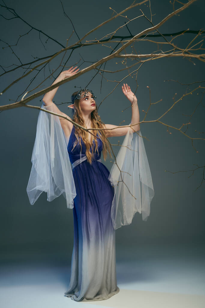 Mladá žena v modrých a bílých šatech drží jemnou větev, ztělesňující pohádkovou princeznu v rozmarném prostředí. - Fotografie, Obrázek