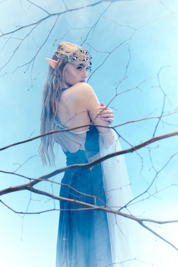 Молодая женщина в синем платье, похожая на принцессу эльфов, грациозно стоит перед величественным деревом. - Фото, изображение