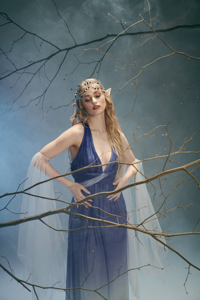 Une jeune femme en robe bleue se tient gracieusement devant un arbre majestueux, incarnant une présence féerique dans un décor de studio. - Photo, image