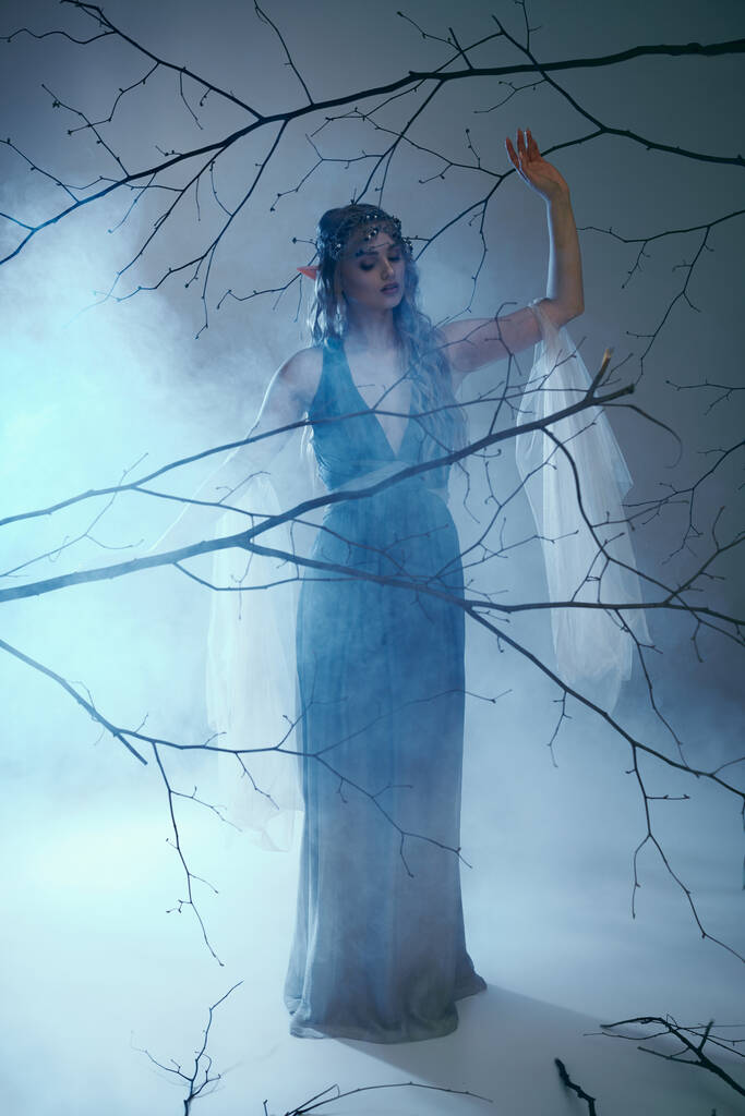Μια νεαρή γυναίκα με ένα μπλε φόρεμα στέκεται με χάρη σε ένα ομιχλώδες περιβάλλον, ενσωματώνοντας την ουσία μιας παραμυθένιας πριγκίπισσας ξωτικών.. - Φωτογραφία, εικόνα