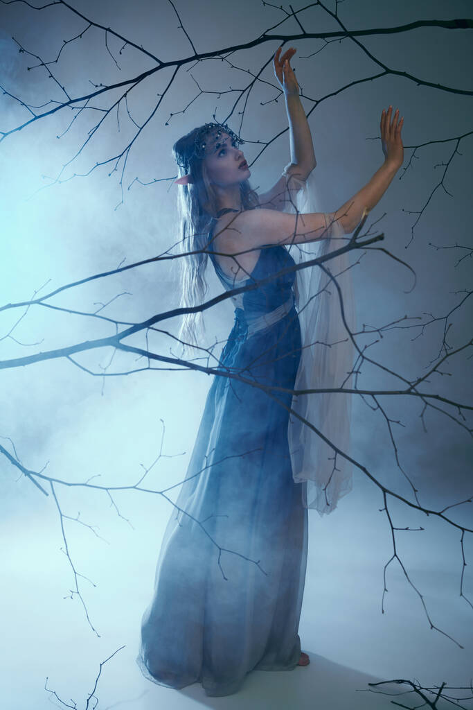 Μια νεαρή γυναίκα με μπλε φόρεμα στέκεται με χάρη μπροστά σε ένα μεγαλοπρεπές δέντρο, ενσαρκώνοντας την ουσία μιας αιθέριας πριγκίπισσας ξωτικών.. - Φωτογραφία, εικόνα
