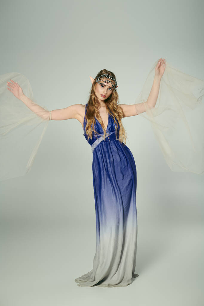 Μια νεαρή γυναίκα στέκεται κομψά σε ένα σκηνικό στούντιο, ντυμένη με ένα όμορφο μπλε και γκρι φόρεμα κατάλληλο για μια πριγκίπισσα παραμύθι. - Φωτογραφία, εικόνα