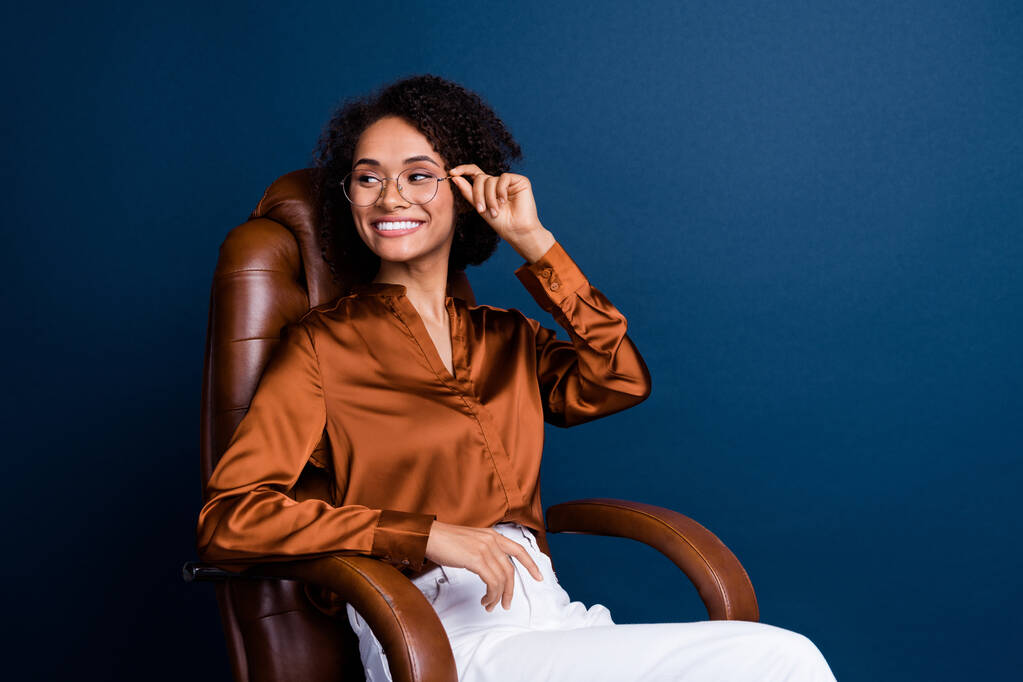 美しい若い女性のオフィスの家具の広告の写真の肖像画は暗い青い背景で隔離された流行の茶色のサテン フォーマルウェアを着ます. - 写真・画像